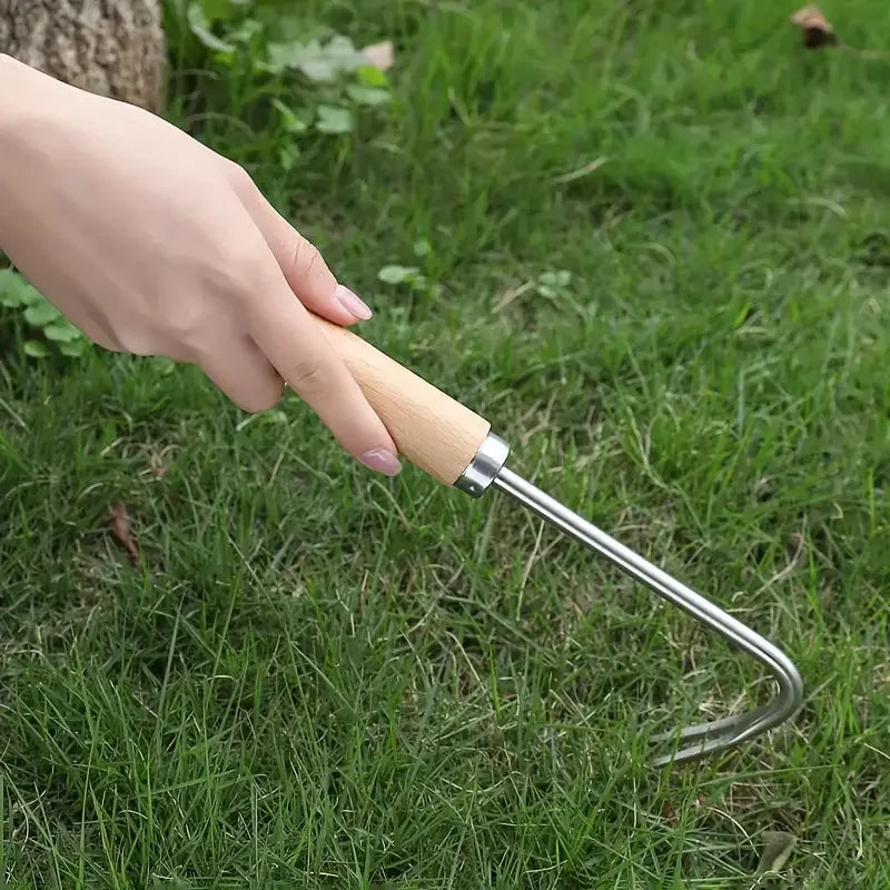 A hand holds a stainless steel garden weeder with a wooden handle in a grassy outdoor setting.