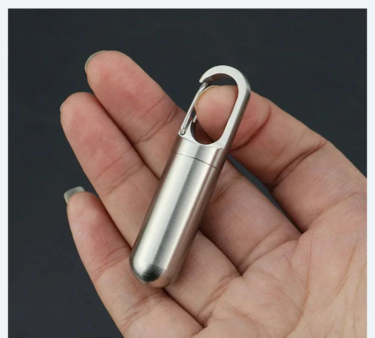 A hand holds a stainless steel waterproof pill capsule with a keychain attached.