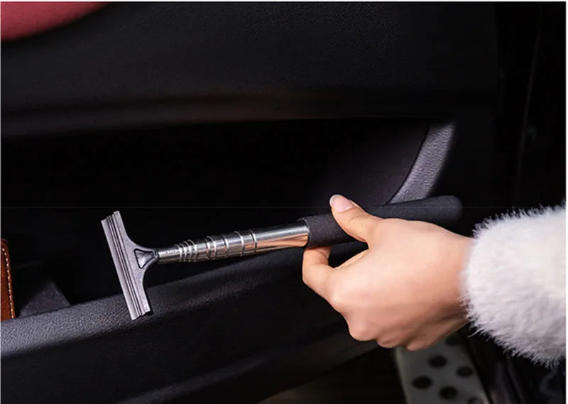 A hand is using a silver retractable car mirror and window cleaning wiper on a black vehicle interior.