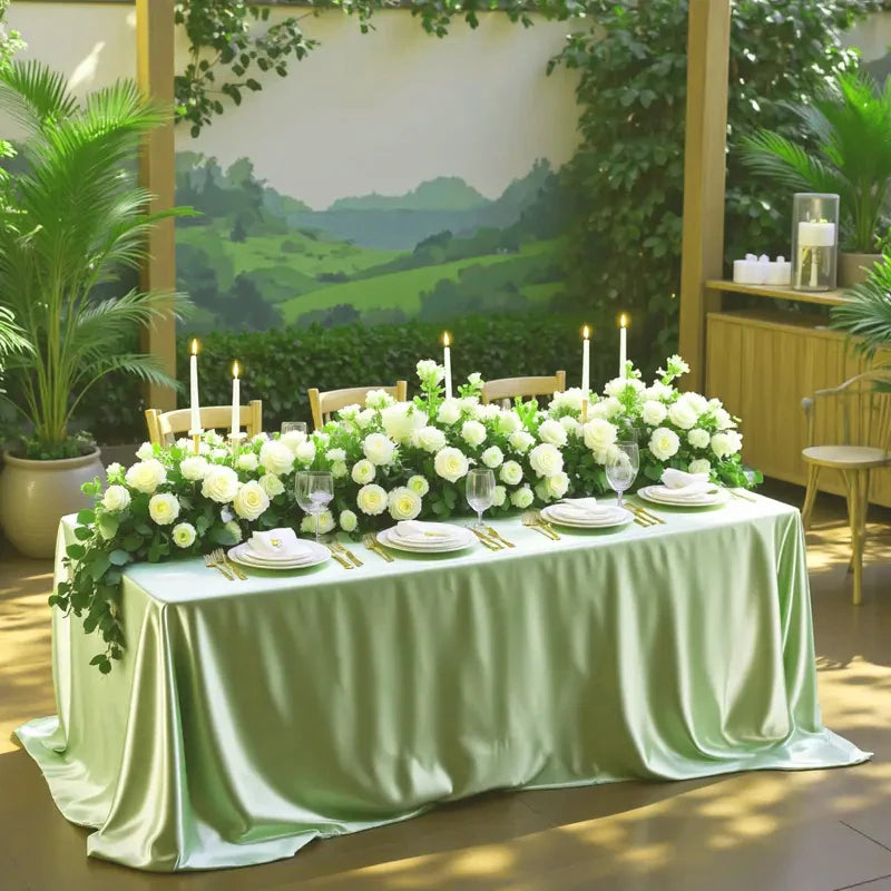 An elegant round satin tablecloth draped in sage green fabric, decorated with white flowers and candles for an event.