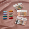 A set of eight classic solid color scarf pins in various shades of green, yellow, and pink are displayed on a pink fabric.