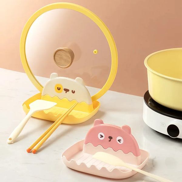 A yellow countertop pot lid and utensil organizer with wooden spoons and chopsticks on a white surface.