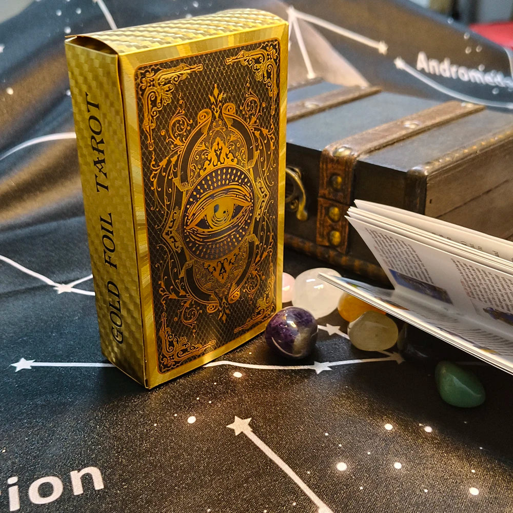 A gold foil tarot card deck box with an eye design rests on a dark surface next to crystals and a guidebook.