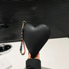 A black heart-shaped PU coin purse with a leather wrist strap is held up against a dark background.