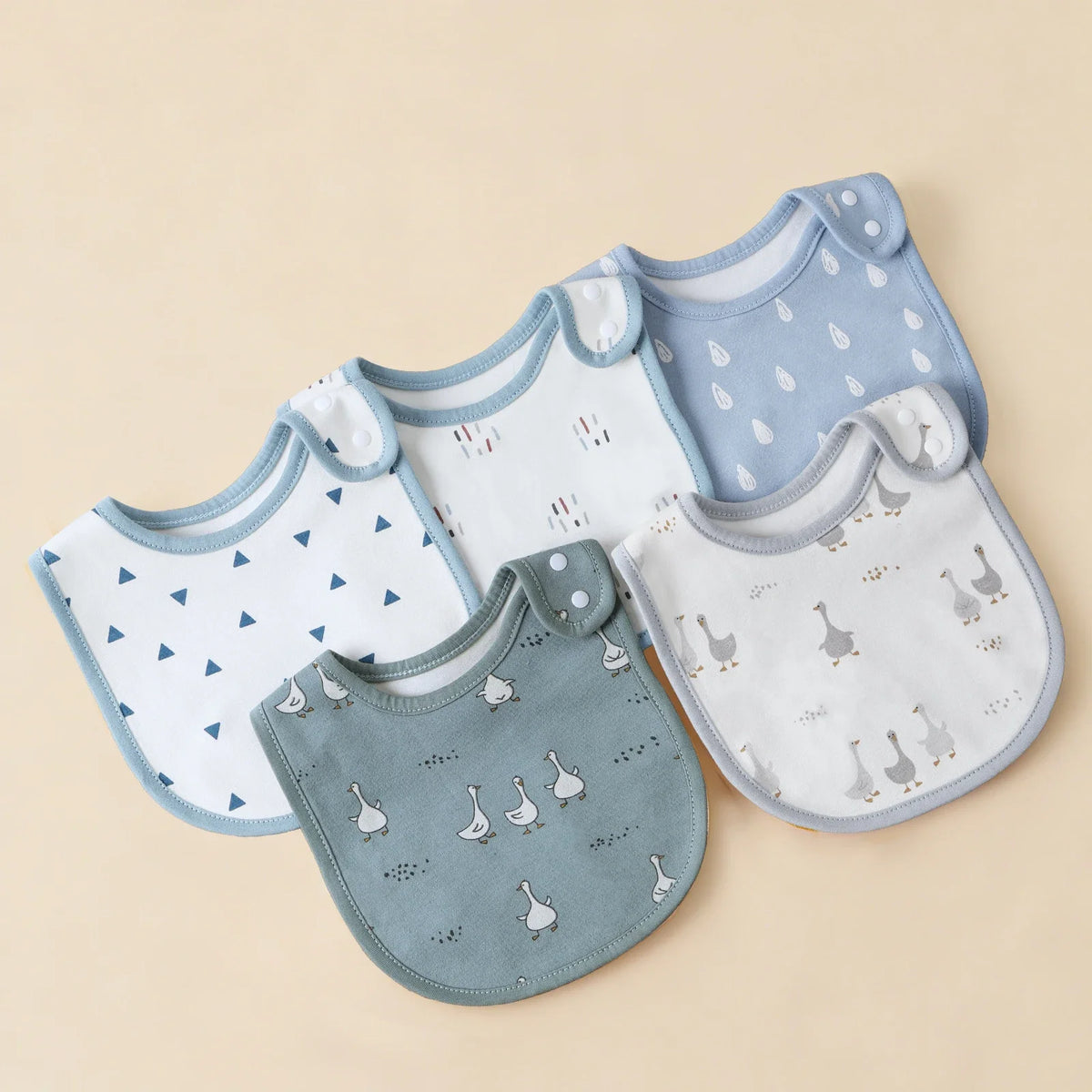 A collection of light blue and gray cotton baby bibs with various patterns and shapes laid out on a beige background.