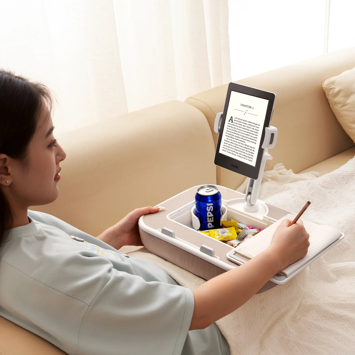 A woman is using a 360° rotatable phone stand and lap desk to view content on a tablet while relaxing on a sofa.