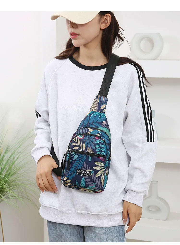 A woman wearing a white sweatshirt holds a blue and green leaf print sling crossbody bag.