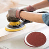 A hand wearing a glove is using an orbital sander to smooth a piece of wood with sanding discs.