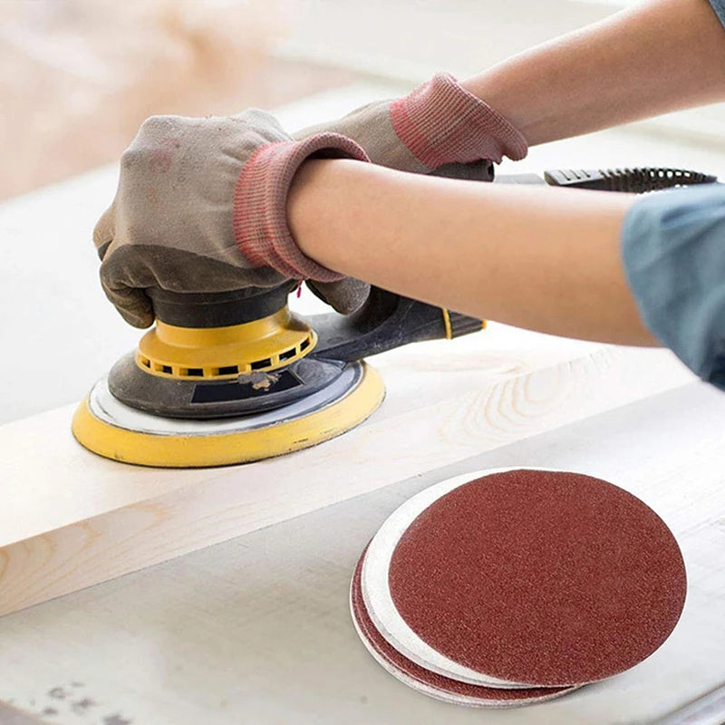 A hand wearing a glove is using an orbital sander to smooth a piece of wood with sanding discs.