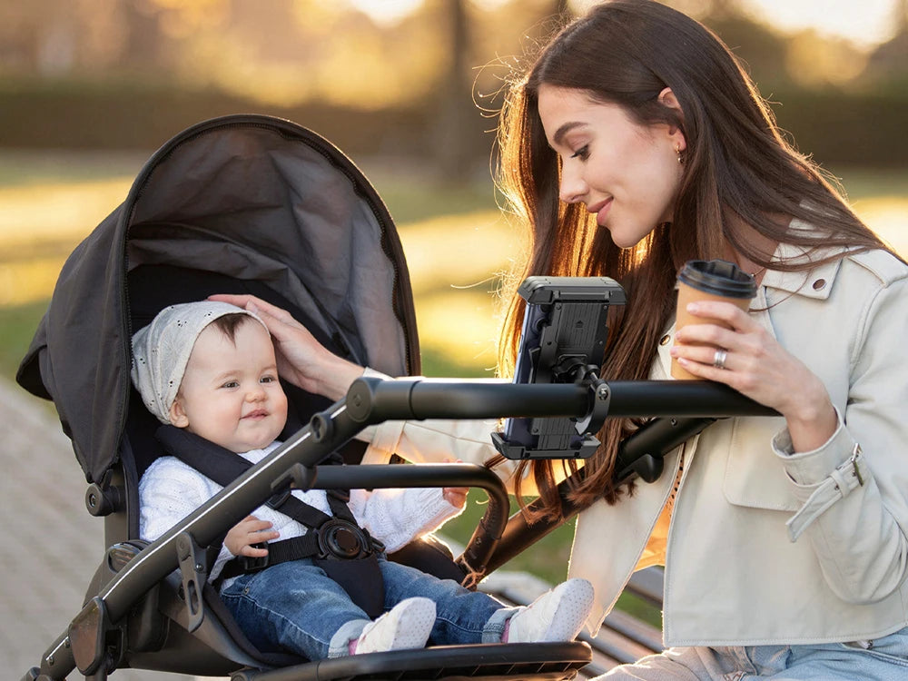 Eine Frau hält ein Baby in einem Fahrrad-Handyhalter mit verstecktem Tracker vor einem weichen Outdoor-Hintergrund.