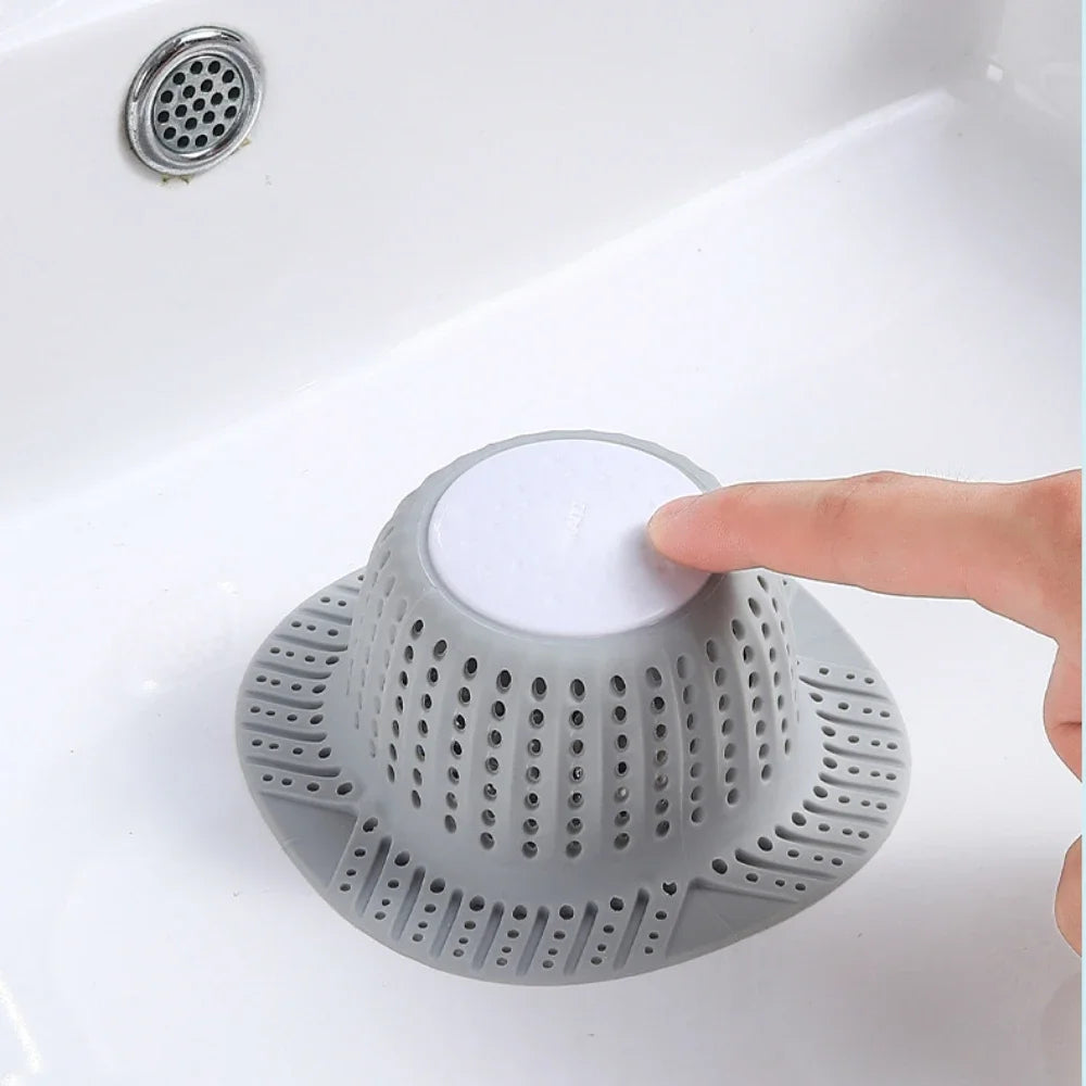A hand is placing a white silicone drain hair catcher into a grey mesh strainer in a white sink.