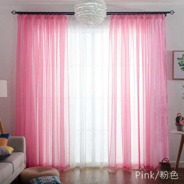 Pink sheer voile curtains with a white pompom hanging in a bright window