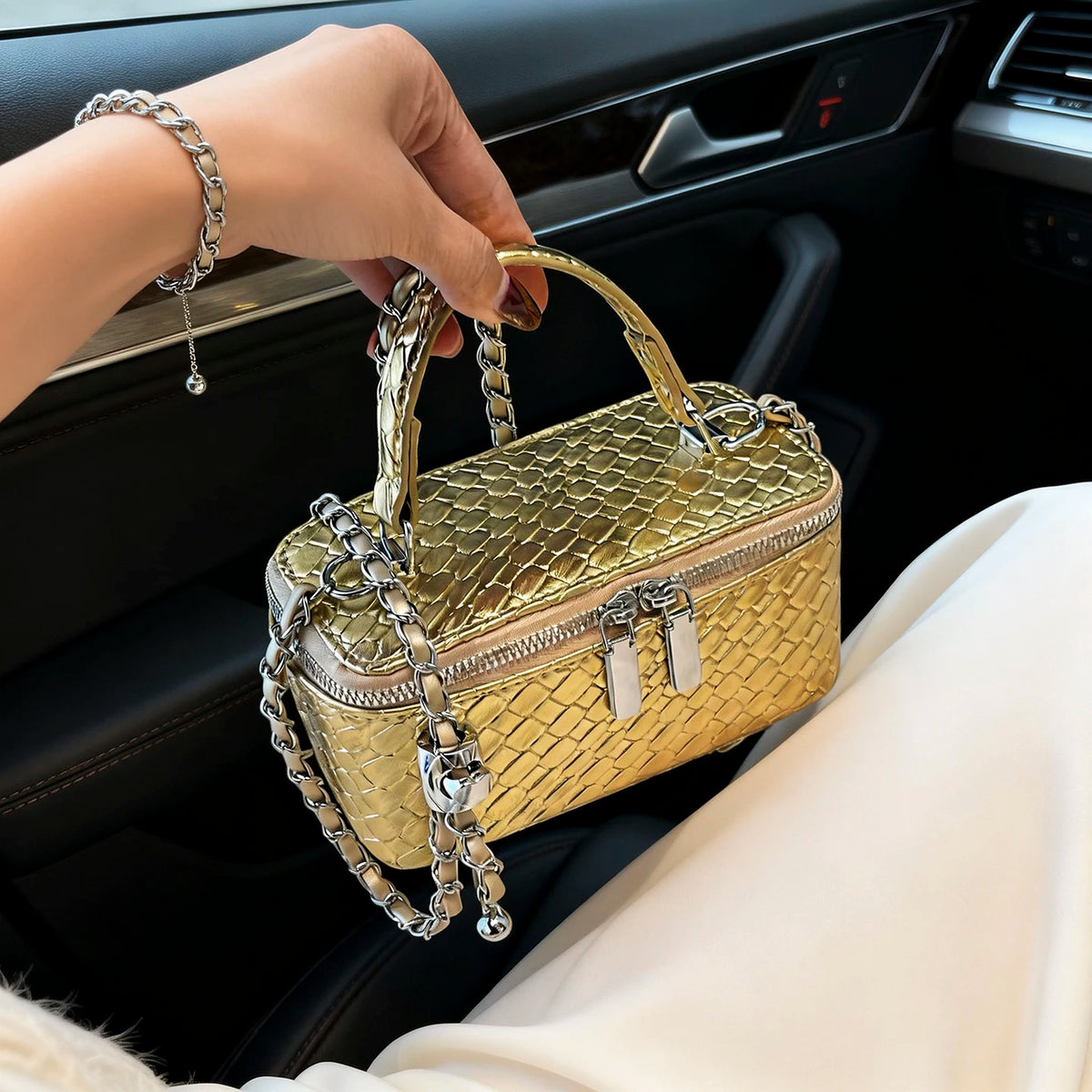 A hand holds a gold woven leather crossbody bag with silver chains inside a car interior.