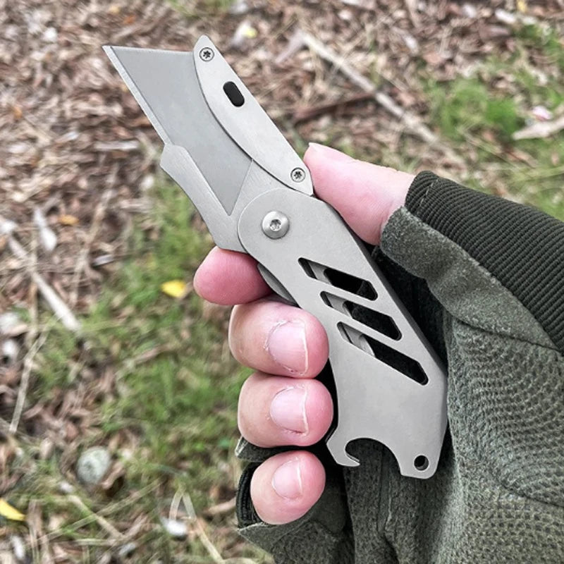 A hand holds a stainless steel folding utility knife with a blade partially open, set against a natural outdoor background.