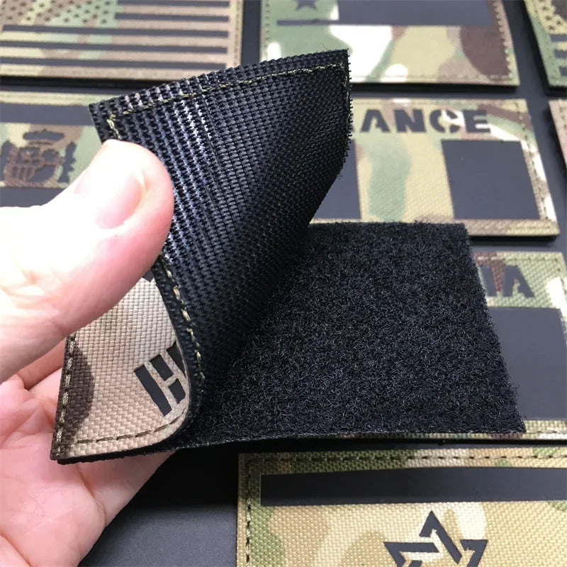 A hand holds a black tactical patch with a beige flag design and hook-and-loop backing against a camouflage background.