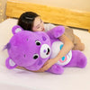 Una mujer abraza un peluche Kawaii de Care Bears arcoíris morado mientras está tumbada en una cama blanca.
