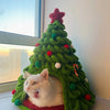 A white cat peeking out of a festive green knitted Christmas tree decorated with red and green ornaments.