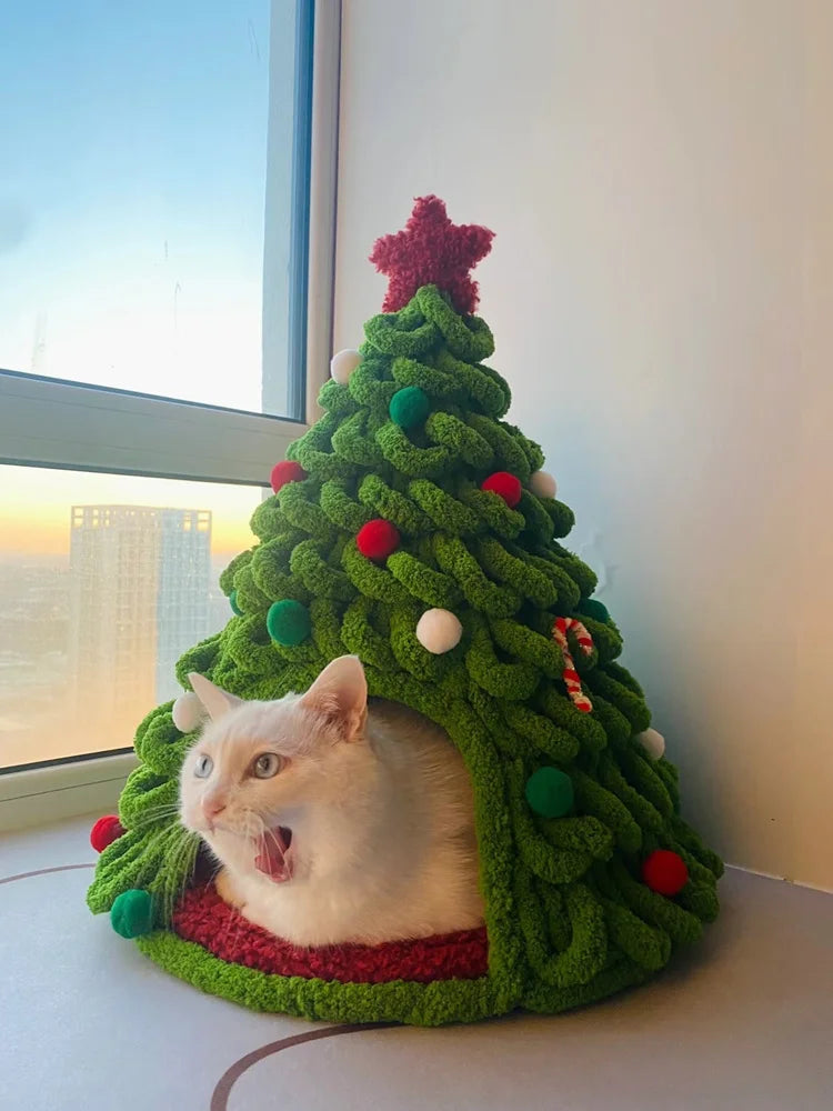 A white cat peeking out of a festive green knitted Christmas tree decorated with red and green ornaments.