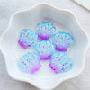 Five iridescent blue and purple resin sea shell charms displayed in a white dish.