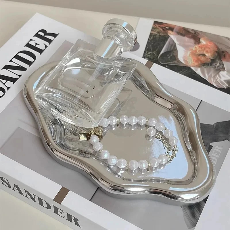 A clear glass perfume bottle and a pearl necklace are displayed on a silver cloud-shaped jewelry tray.
