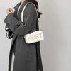 Eine Frau hält eine weiße Leder-Tasche mit goldenen Buchstaben vor einem grauen Mantel.