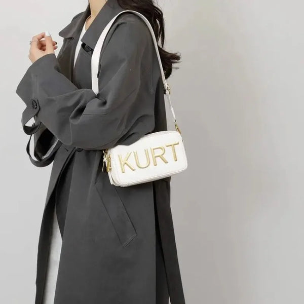 Eine Frau hält eine weiße Leder-Tasche mit goldenen Buchstaben vor einem grauen Mantel.