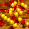 A collection of heart-shaped yellow LED candles resting on golden fabric with scattered red rose petals.