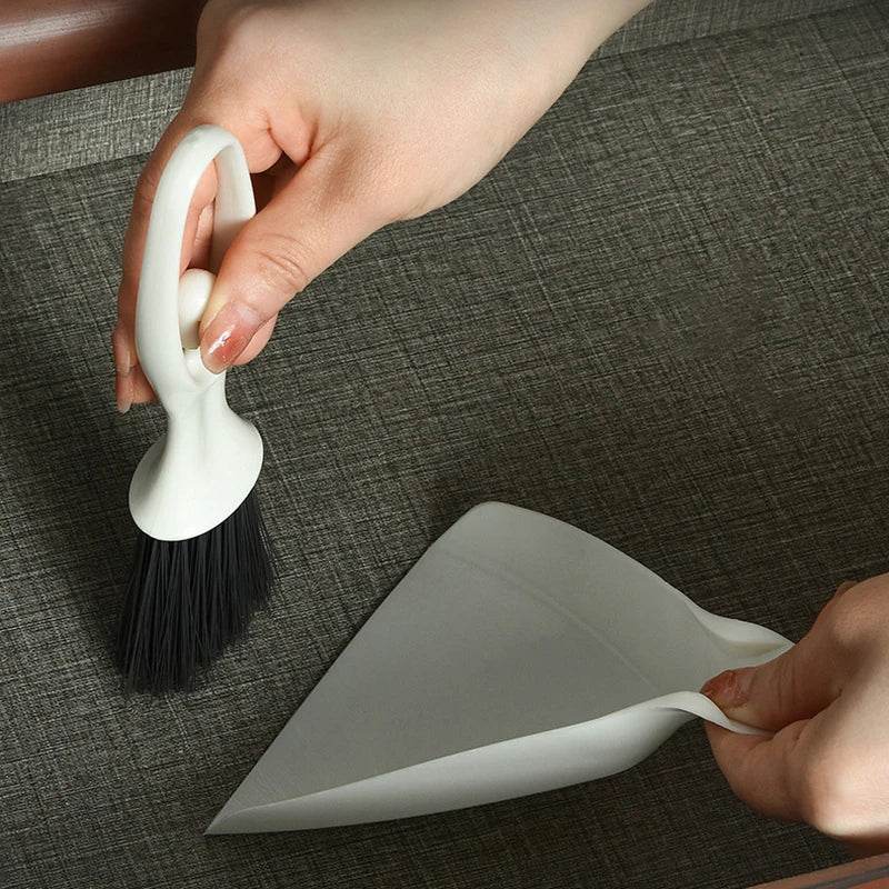 A hand is holding a white compact mini broom and dustpan set on a dark surface.