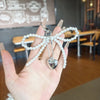 A hand holds a pearl bow keychain with a heart charm against a blurred indoor background.