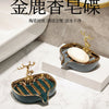 A golden leaf-shaped ceramic soap dish with a drain rack is displayed on a white marble surface.