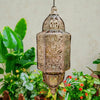 A bronze solar-powered Moroccan lantern with intricate ironwork hanging above lush green foliage.