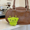 A bright lime green alien plush keychain with small antennae is displayed next to a brown leather handbag.