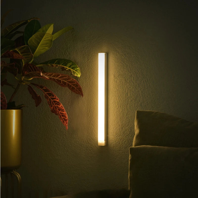 A vertical illuminated night light mounted on a wall next to a potted plant in a dimly lit room.
