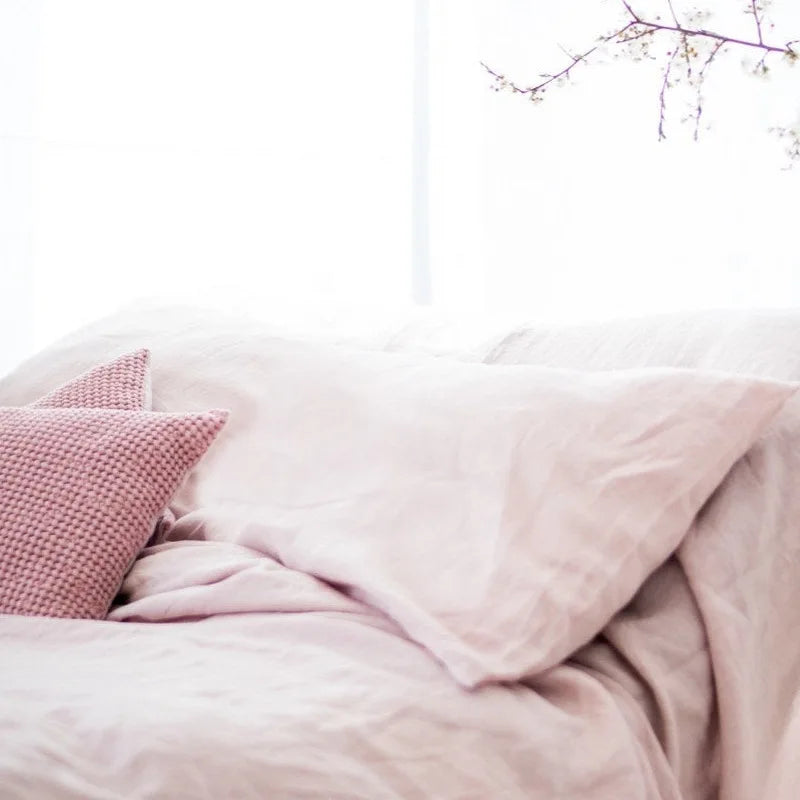 A close-up of a soft pink linen pillowcase with a textured cover and envelope closure on a bed.
