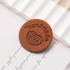 A round, brown PU leather label with the word handmade and a coiled heart design on a white paper background.