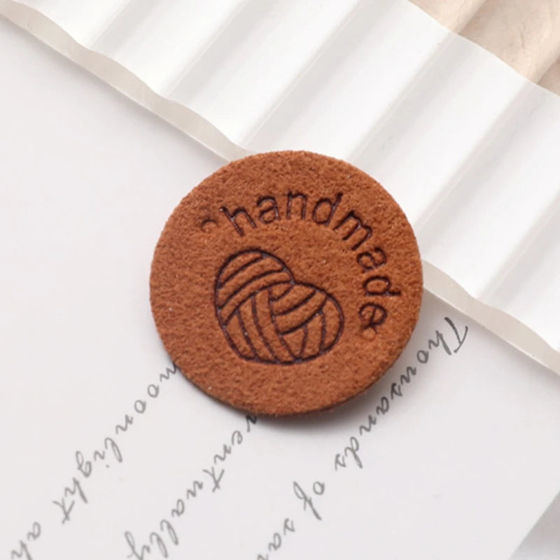 A round, brown PU leather label with the word handmade and a coiled heart design on a white paper background.