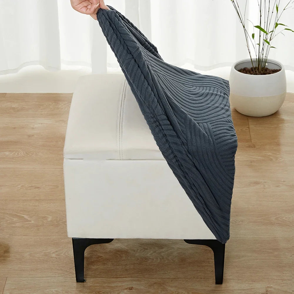 A dark gray geometric jacquard stretch ottoman cover draped over a white upholstered ottoman.