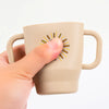 A hand holds a beige silicone training cup with straw and handles featuring a sunburst design.