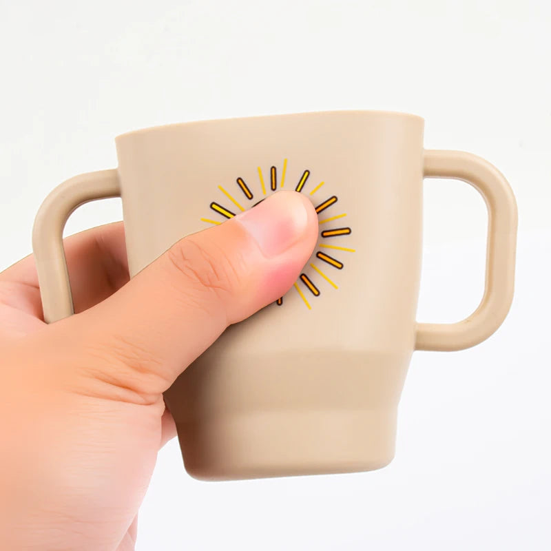A hand holds a beige silicone training cup with straw and handles featuring a sunburst design.