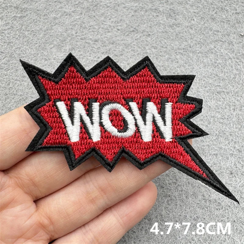A hand holds a red and black embroidered patch shaped like a speech bubble with the word WOW on it.
