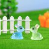 A set of two cute resin miniature bunny figurines, one blue and one green, sitting on bright green grass near a white picket 