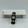 A black fingerboard skateboard is displayed on a light wooden ramp set.