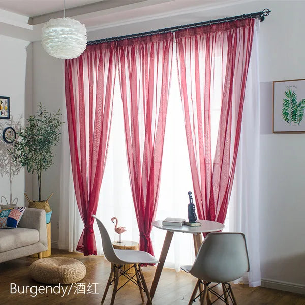 A set of sheer red curtains with a rod pocket filtering light in a bright room.