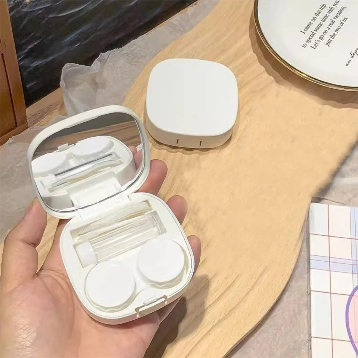 A hand holds an open white compact contact lens travel case with a secure closure and a separate round container.