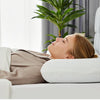 A woman rests her head on a white memory foam pillow in a bright room.