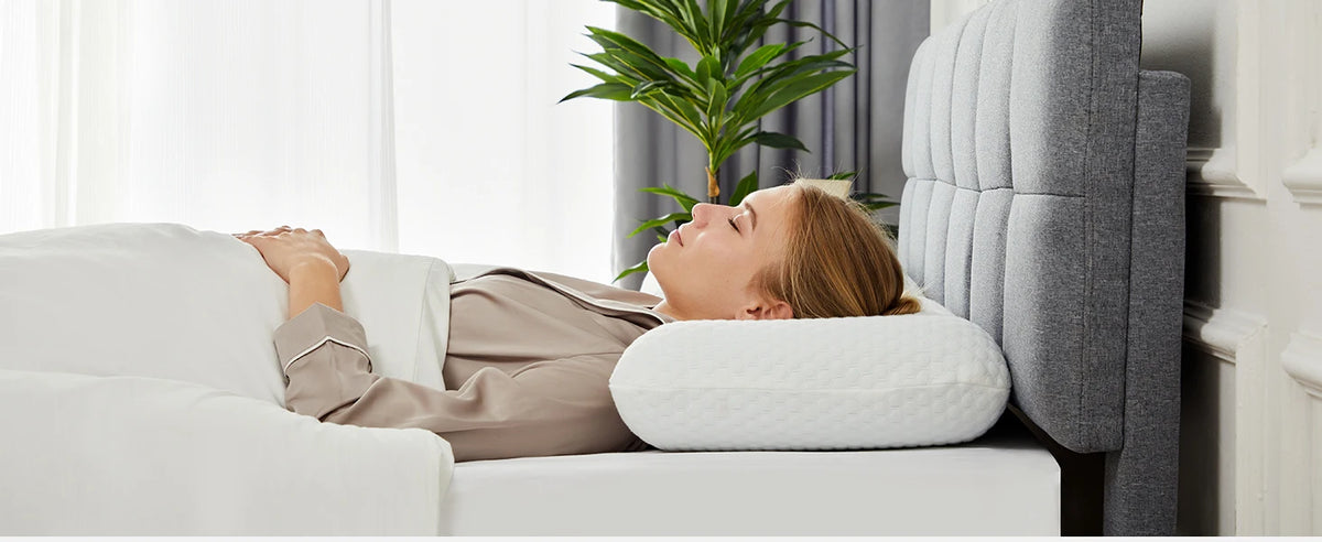A woman rests her head on a white memory foam pillow in a bright room.