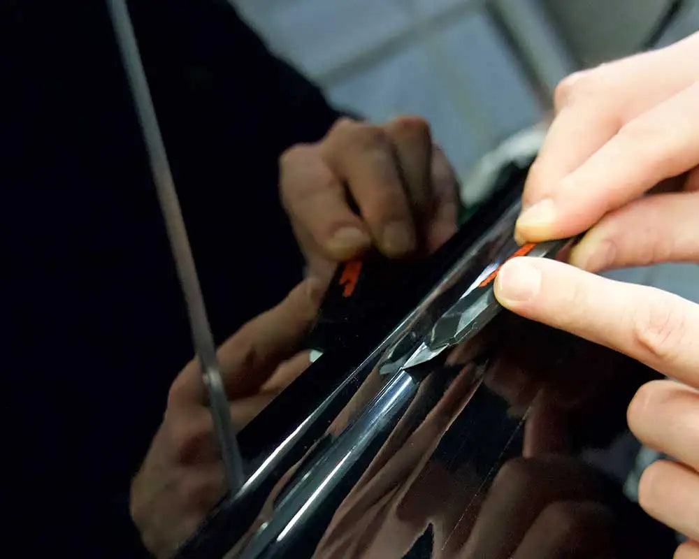 Hands are applying a glossy black vinyl wrap strip to a car trim piece.