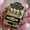 A gold-colored mini fruit slot machine keychain fidget toy with a black display and a black plunger is held in a hand.