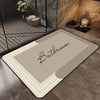 A rectangular beige bath mat with a delicate script reading Bathroom is placed on a tiled floor near a glass shelf.