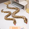 A realistic, soft plush toy of a patterned snake, likely an anaconda doll, lying on a white surface.